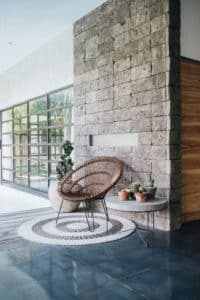 Espace intérieur lumineux de style biophilique, avec mur en pierre naturelle, fauteuil en rotin, table ronde ornée de plantes en pot, sol en béton poli et grande baie vitrée donnant sur un jardin verdoyant.